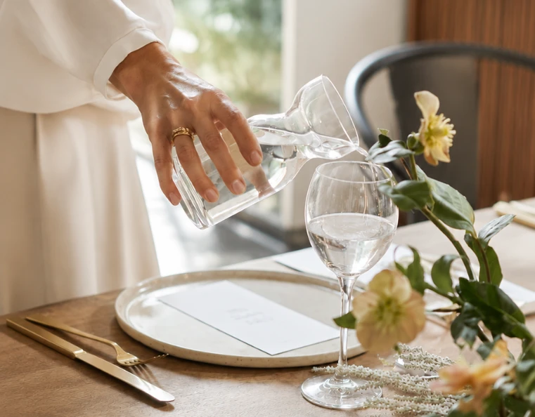 Hand pouring water into a wine glass on an elegant dining table during a luxury hospitality event by Crownpath Hospitality Services in Ermionida Greece, featuring flowers and gold cutlery.