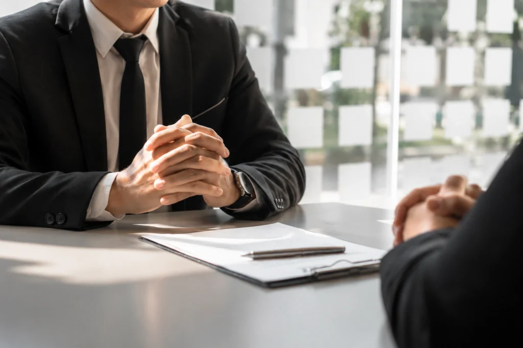 Professional office interview scene with two businesspeople seated across a desk in a bright modern workspace, hands clasped during a formal meeting.